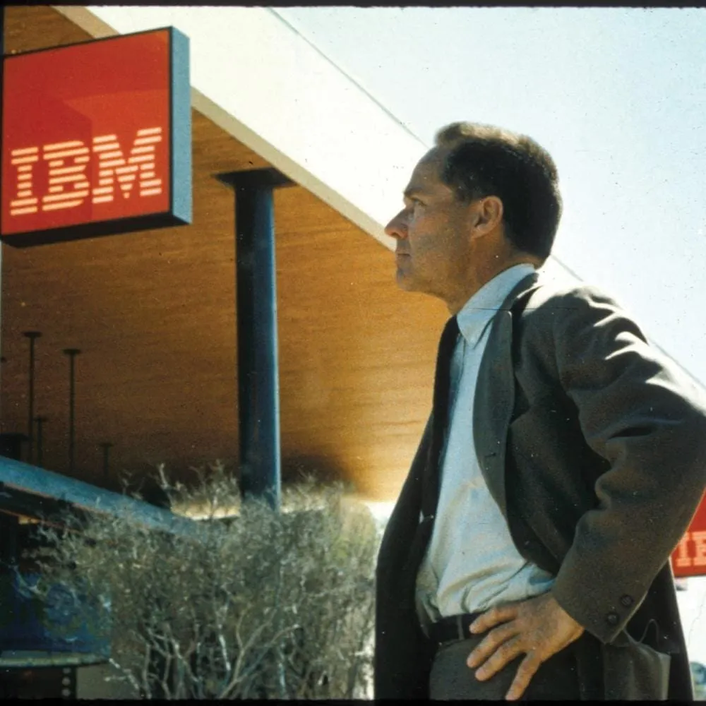 Eliot Noyes de profil, en costume, devant un bâtiment IBM à l’enseigne rouge.
